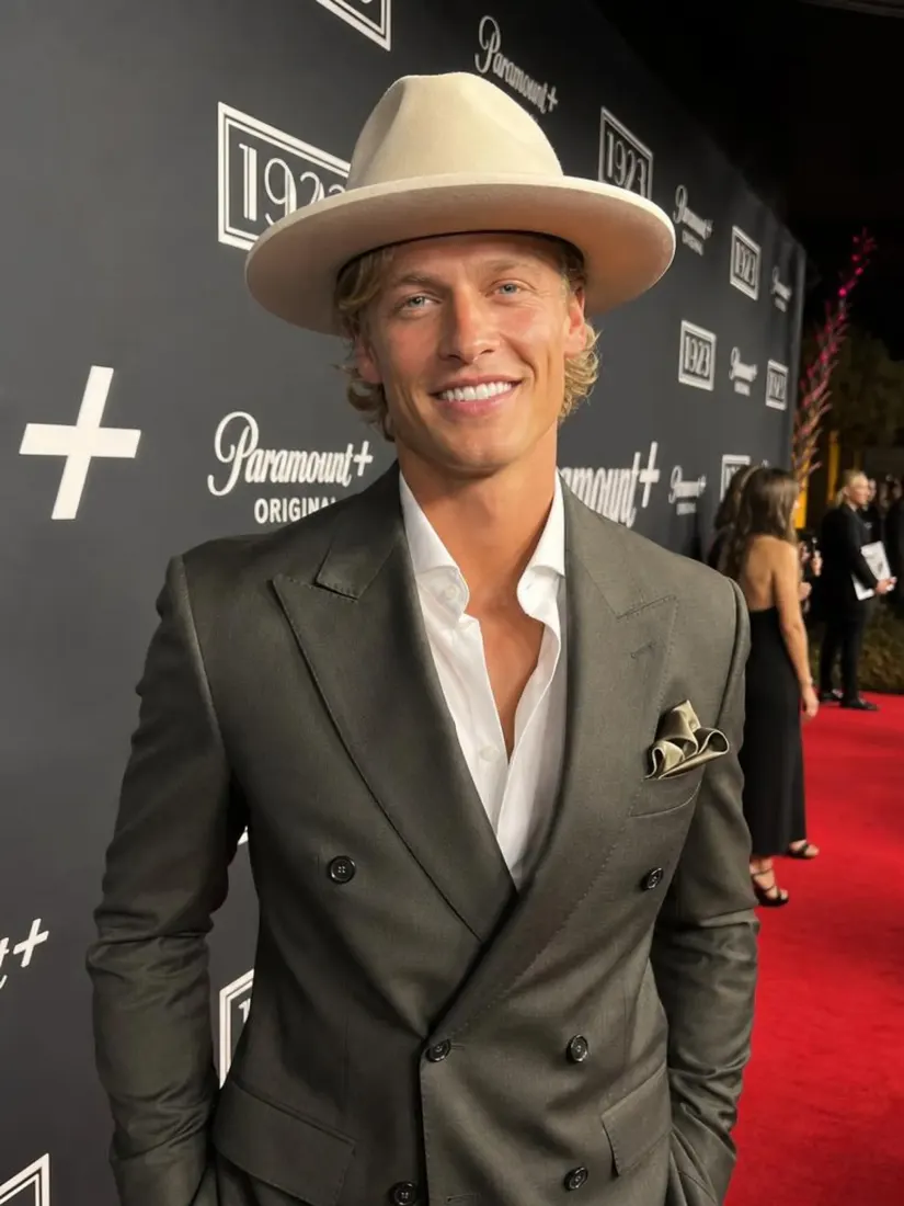 A smiling man in a cream fedora and gray double-breasted suit stands on a red carpet, before the Custom Giant Doll Filter restyles him.