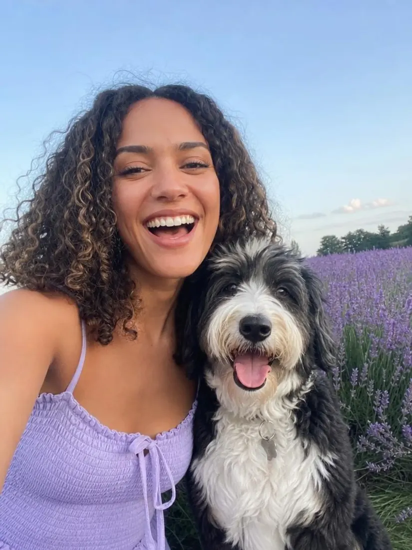 A curly-haired woman laughs while holding a black and white dog in a lavender field, before becoming an FPS game avatar.