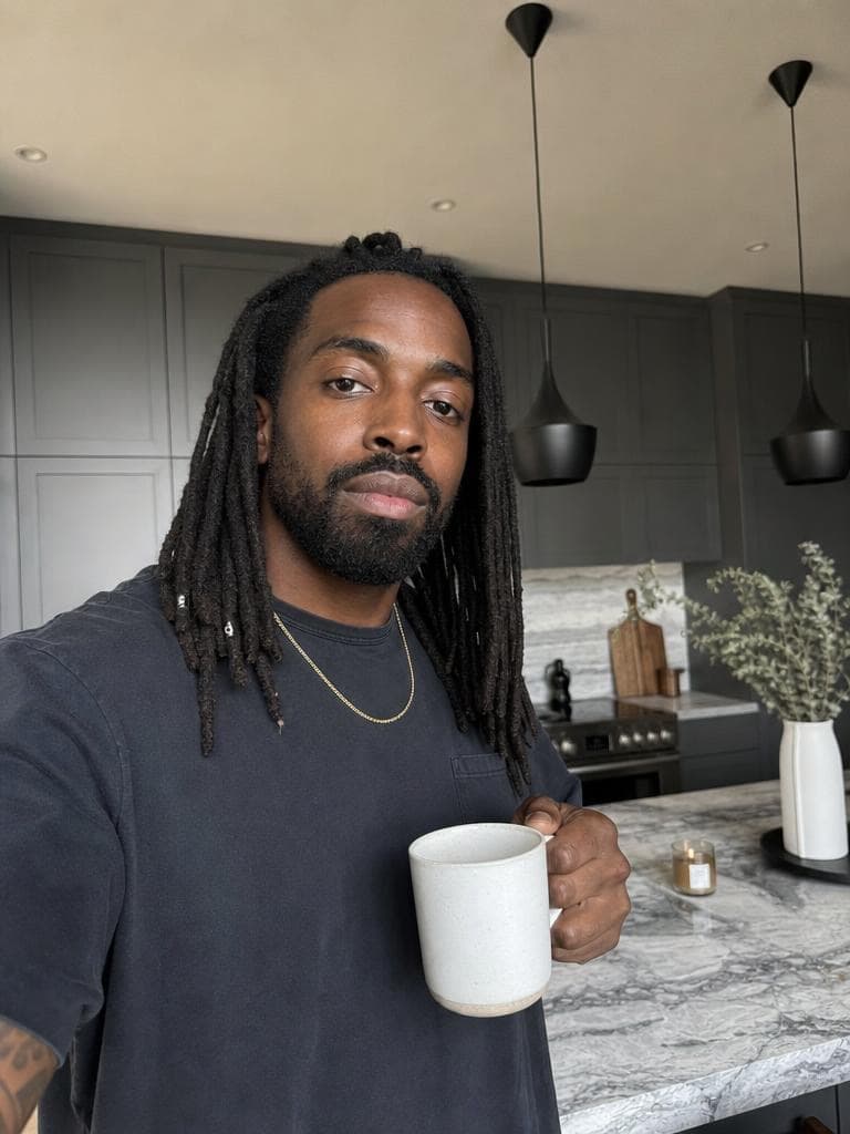 A man with locs holds a mug in a gray kitchen, before the Live Broadcast Generator restyles the scene.